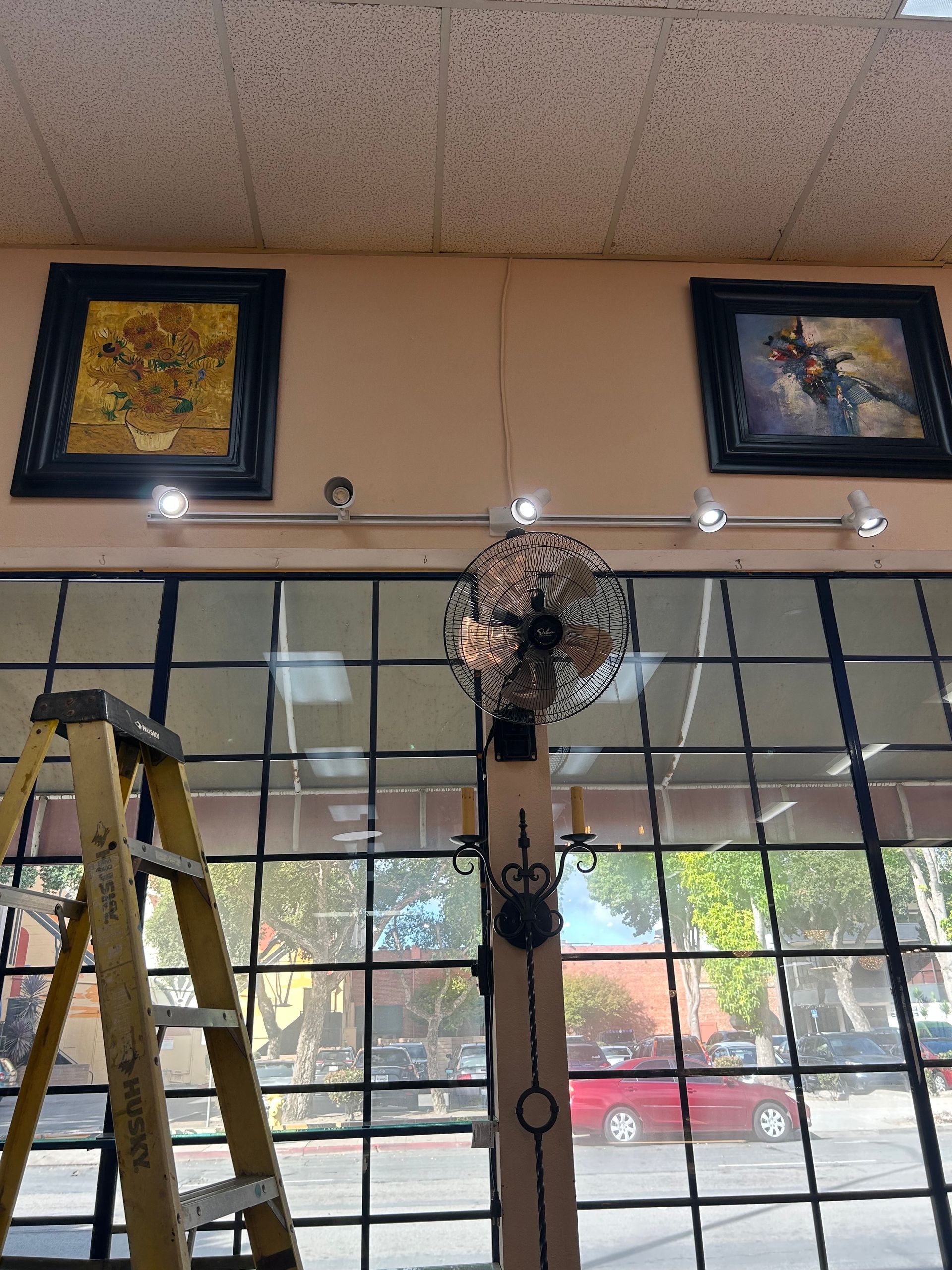 Ladder next to a window with art, lights, and a fan; a street scene is reflected.
