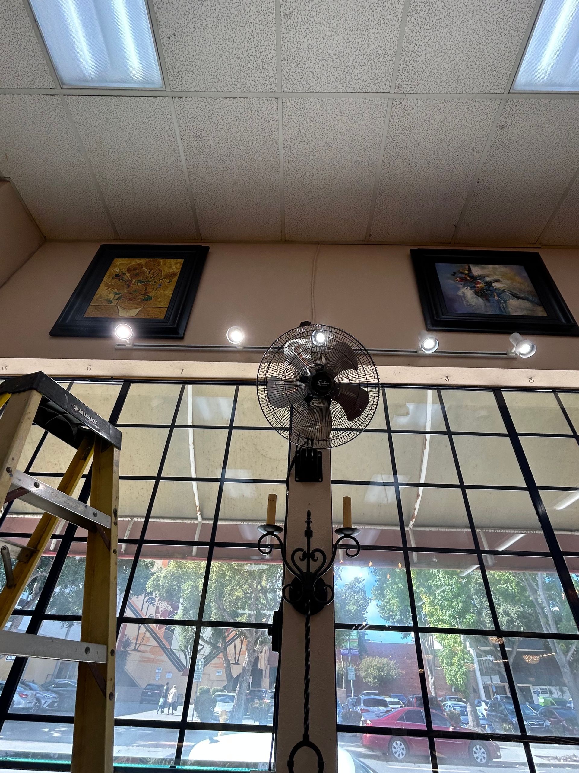 A decorative fan hangs in front of a window. Two framed pictures and a ladder are visible.