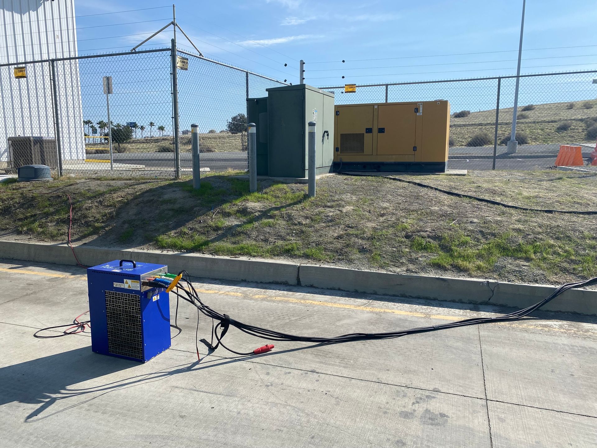 Blue box with wires connected to a generator and electrical box behind a fence on a sunny day.