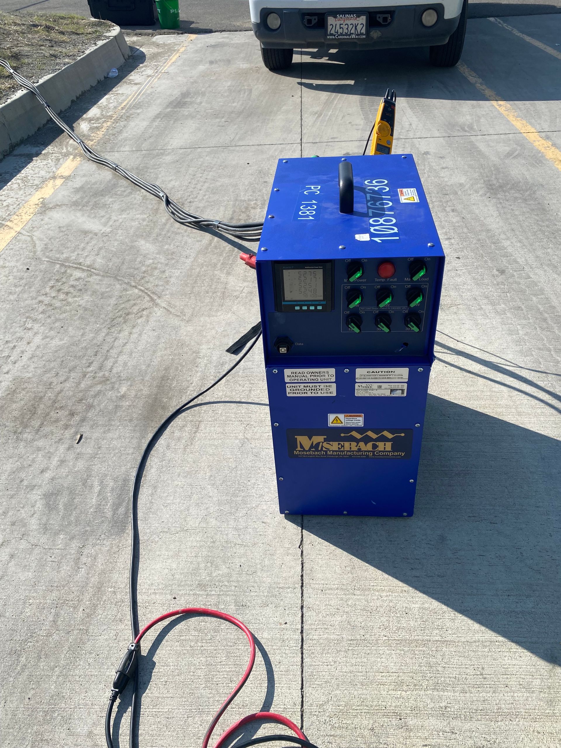 Blue industrial machine on concrete, with cables extending. A white truck is in the background.