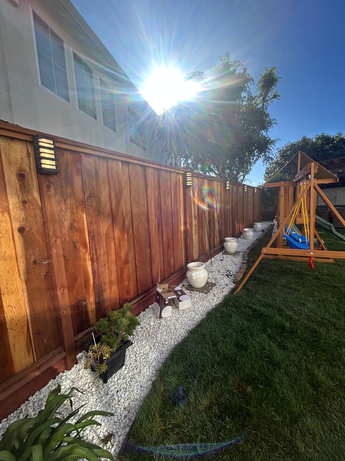 Wooden fence with white gravel path, potted plants, and swing set in sunny backyard.