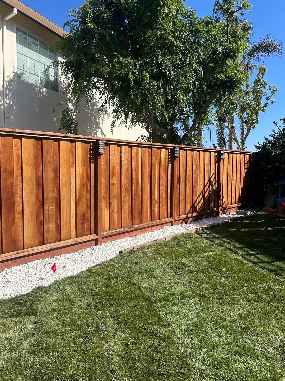 A wood fence in a backyard with green grass, white stones, and a house in the background.