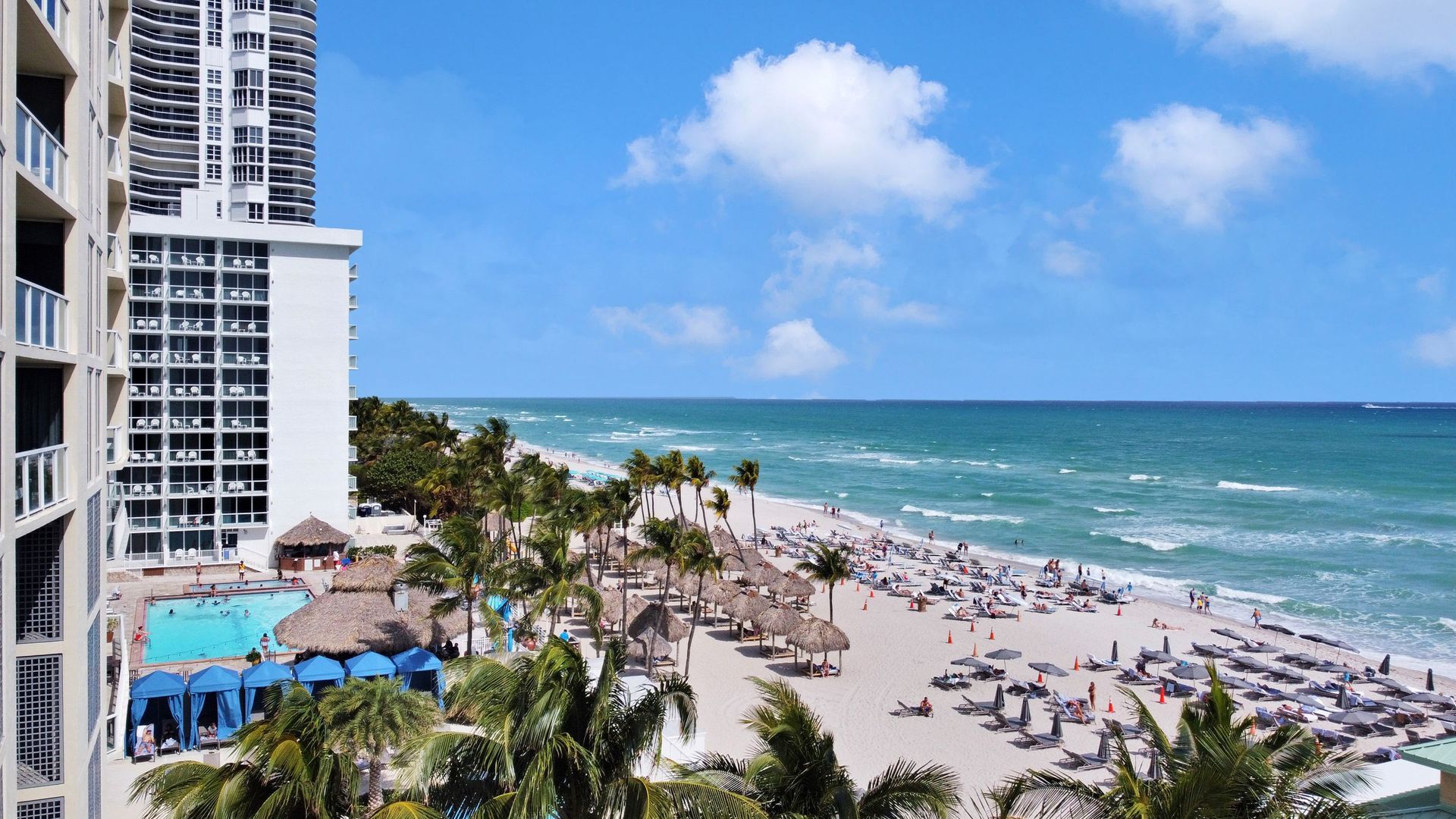 A packed Miami Beach shoreline from above, hotel towers and full rows of umbrellas in peak season