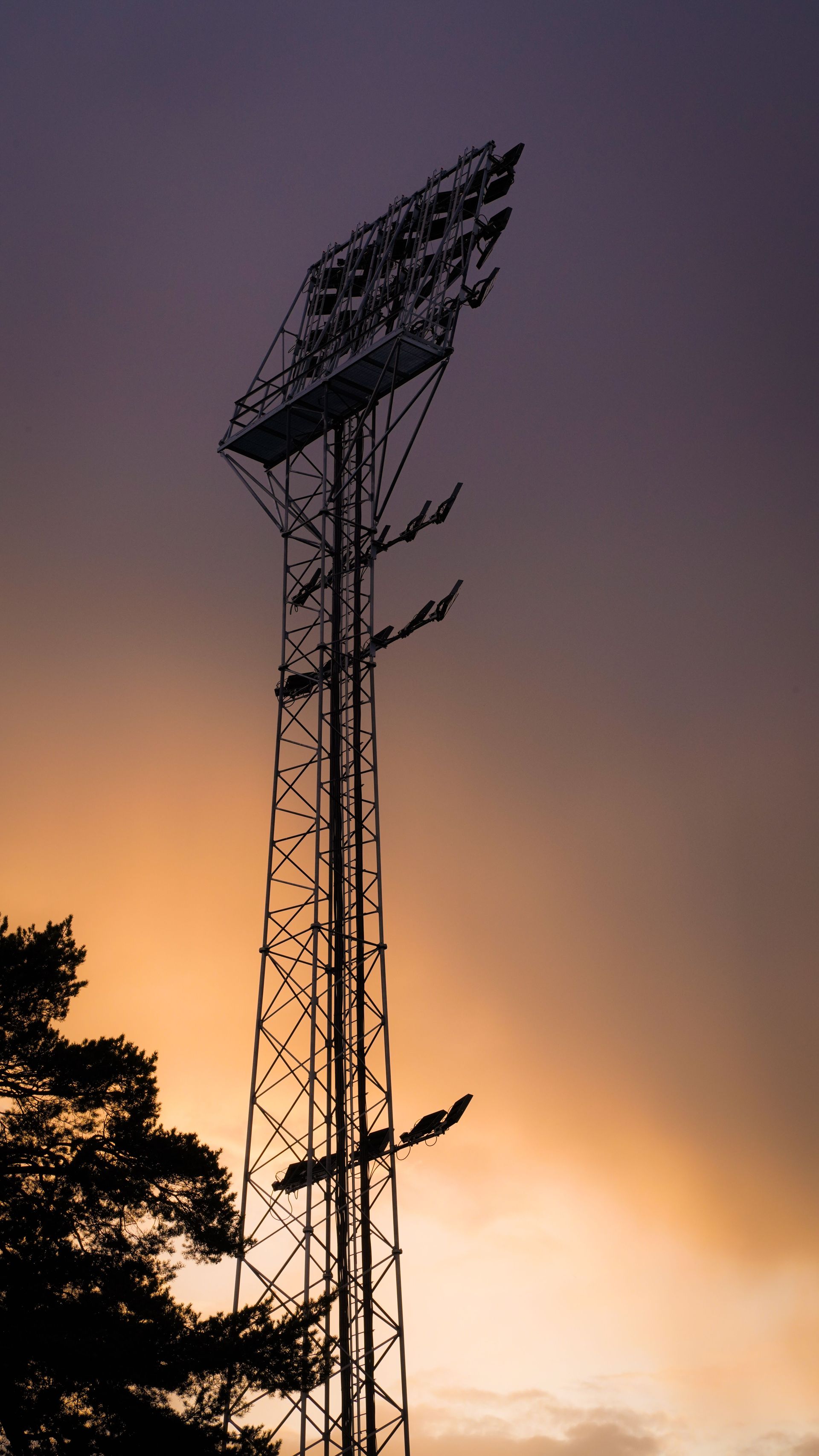 Silhouette of a stadium floodlight tower against an orange sunset sky