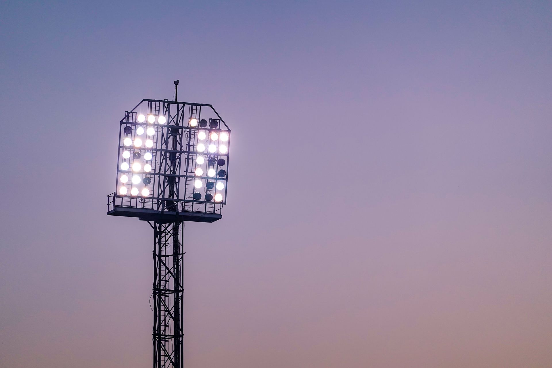 Stadium floodlight tower glowing against a lilac dusk sky