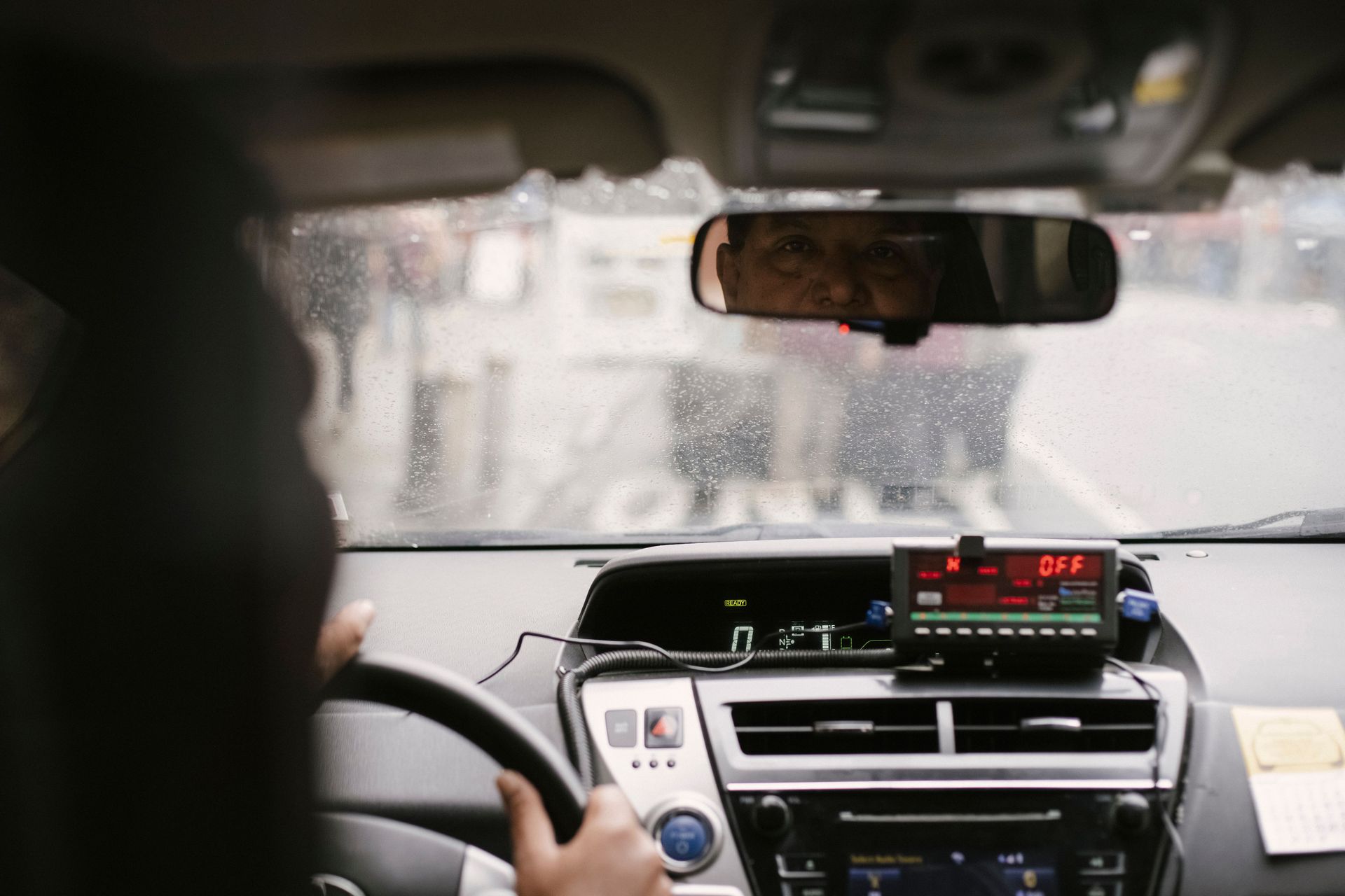 A taxi meter mounted on the dashboard of a car, seen from the back seat through a rain-streaked wind