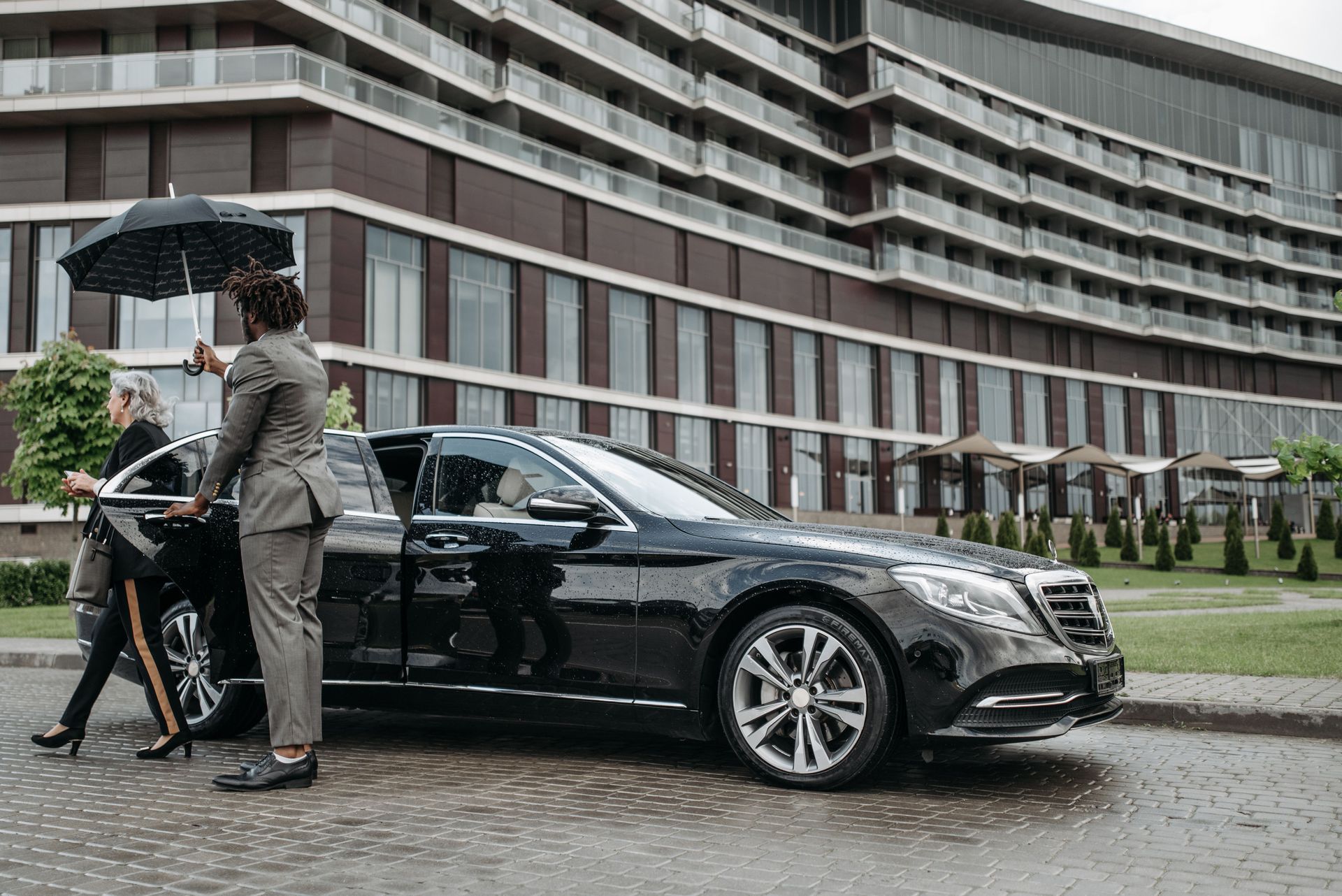 A chauffeur holding an umbrella and opening the rear door of a black Mercedes-Benz S-Class outside a