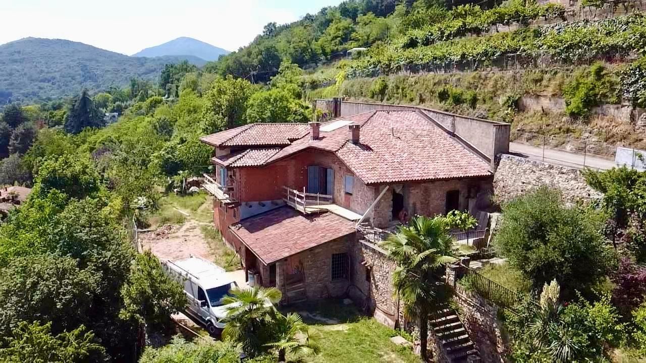 Rustica casa in collina con tetto di tegole rosse, circondata da rigogliosi alberi verdi e montagne.