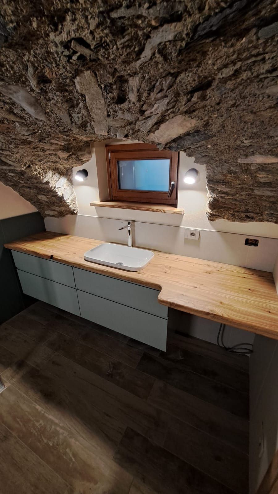 Accogliente bagno mansardato con piano in legno, lavabo, specchio e applique sotto un soffitto inclinato in pietra.