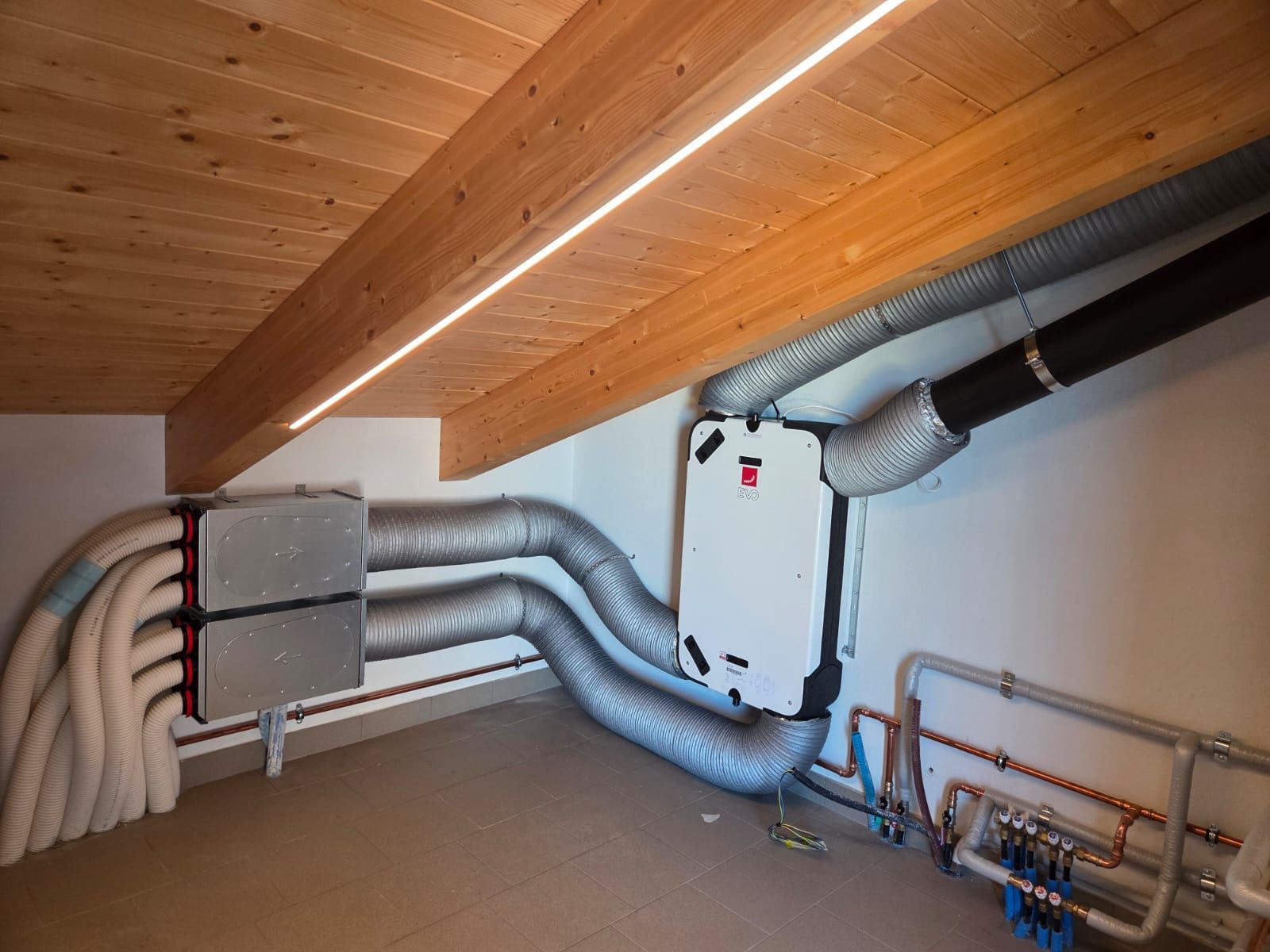 Locale tecnico in mansarda con tubi e condotti isolati e un'unità a parete bianca sotto un soffitto in legno inclinato.