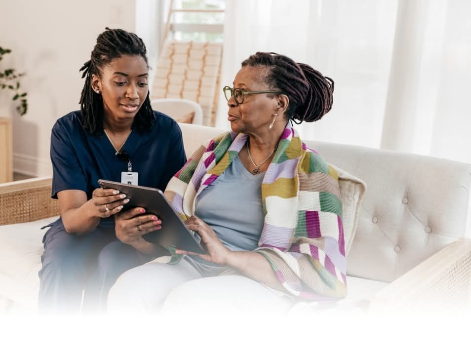 A nurse is sitting next to an older woman on a couch looking at a tablet.