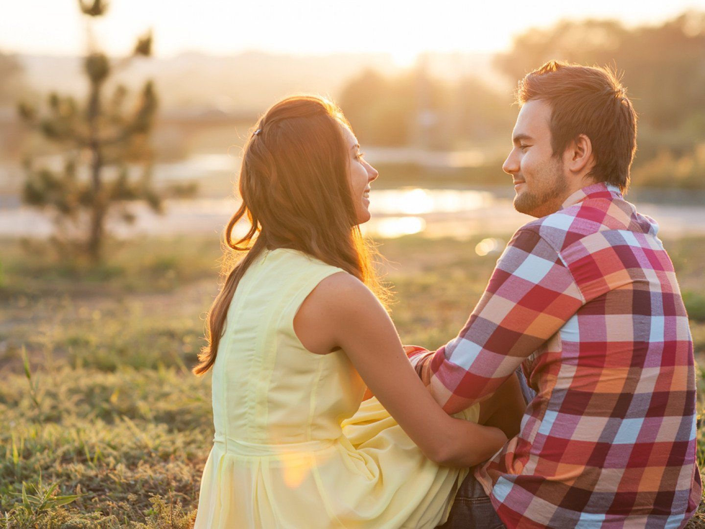 Woman and man smiling, looking at each other outdoors, sunny background.