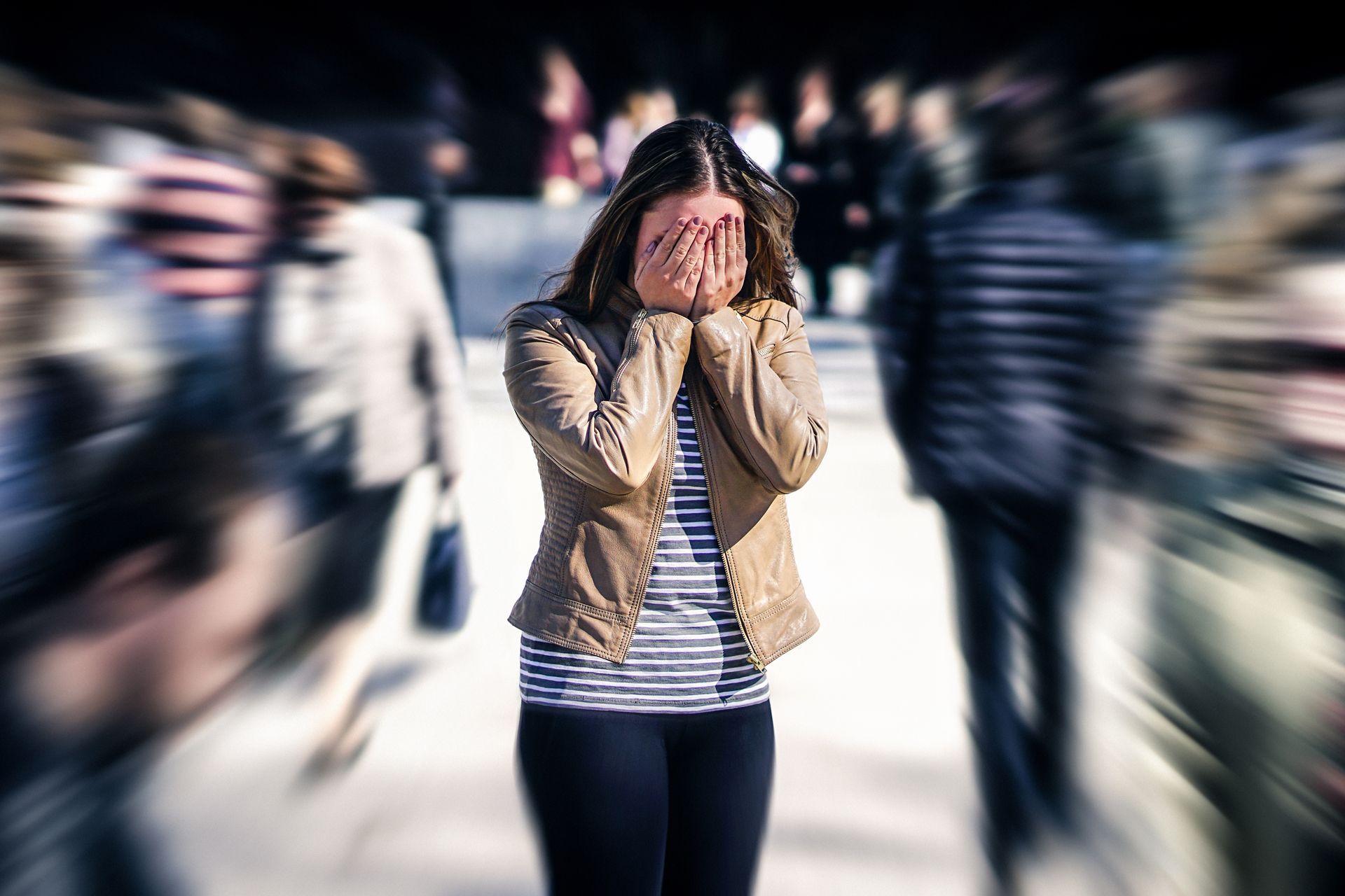 Woman with hands over face, overwhelmed by a blurry crowd in an outdoor setting.