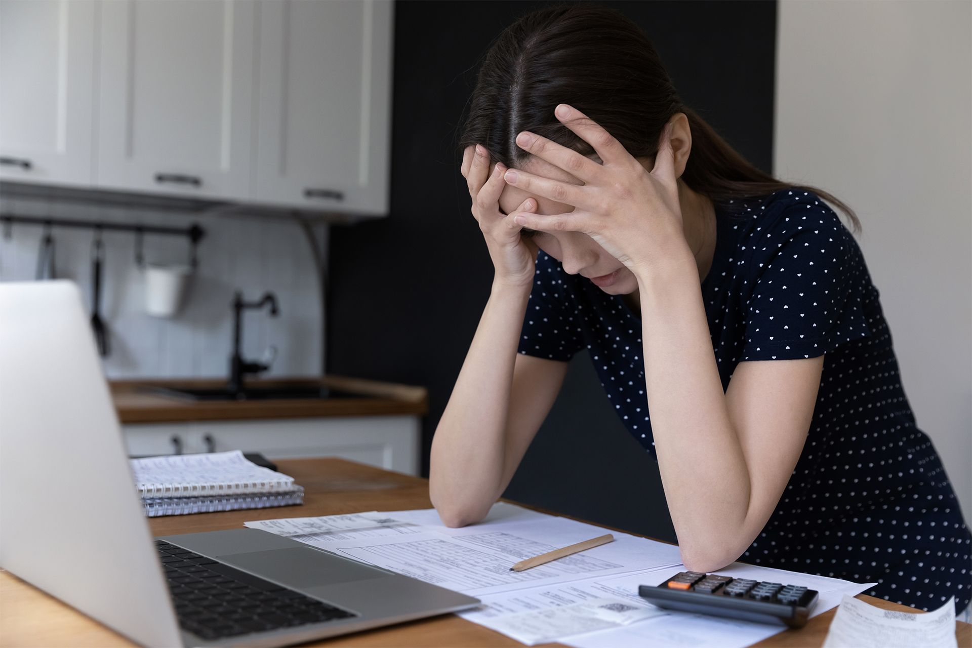 Woman with hands on head, looking at bills and laptop on a table, appearing stressed.