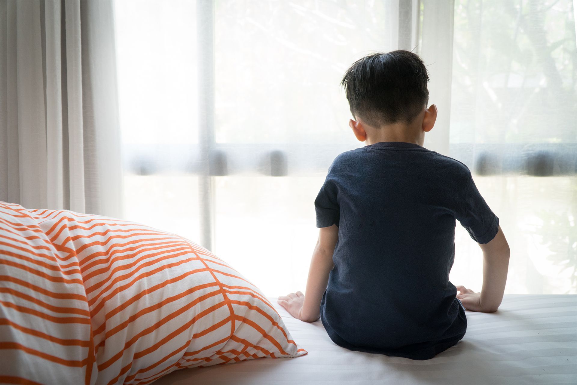 Child sitting on a bed, looking out a window.