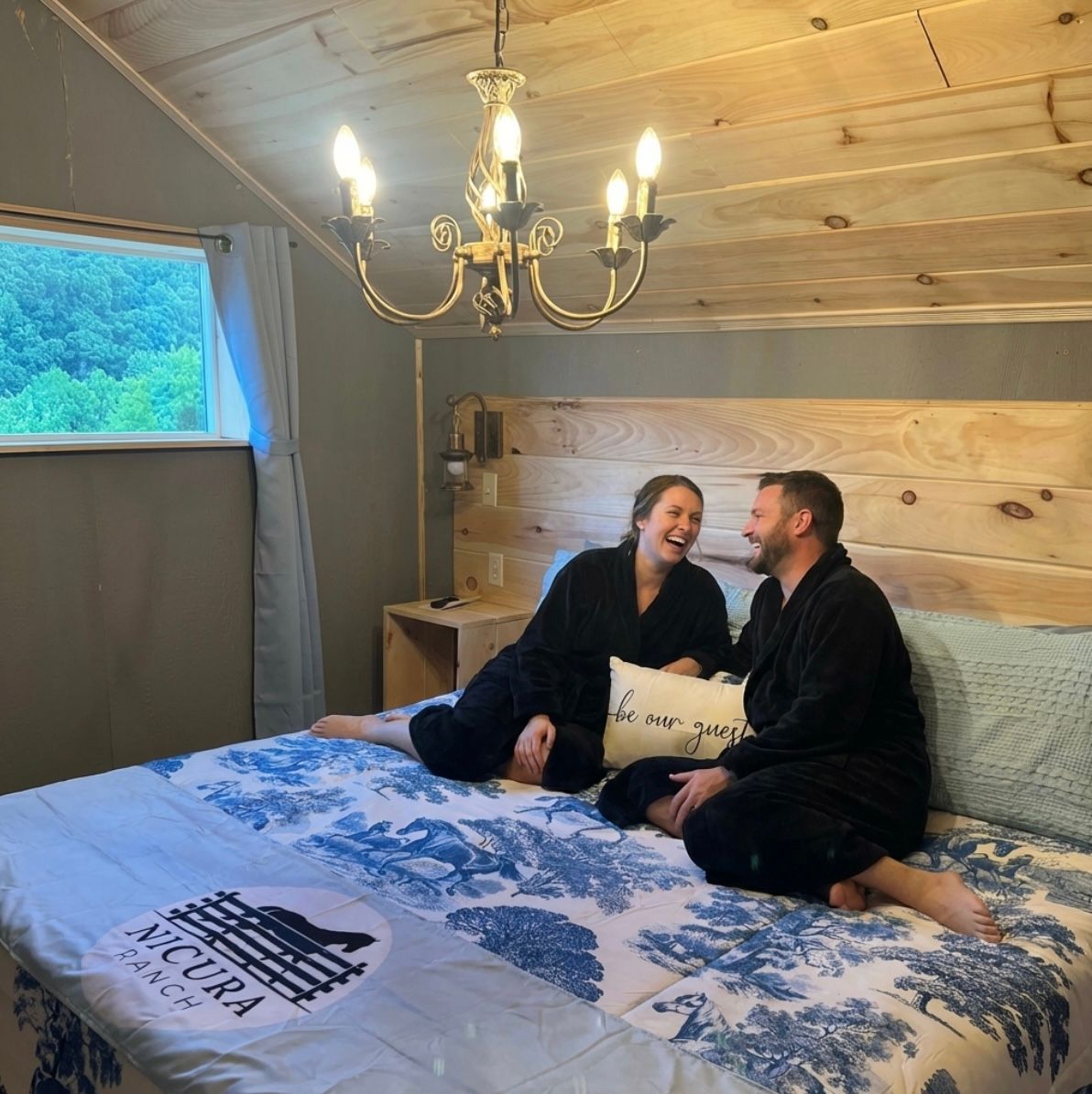Couple laughing on a bed in a cozy wooden loft bedroom with blue patterned bedding and chandelier light