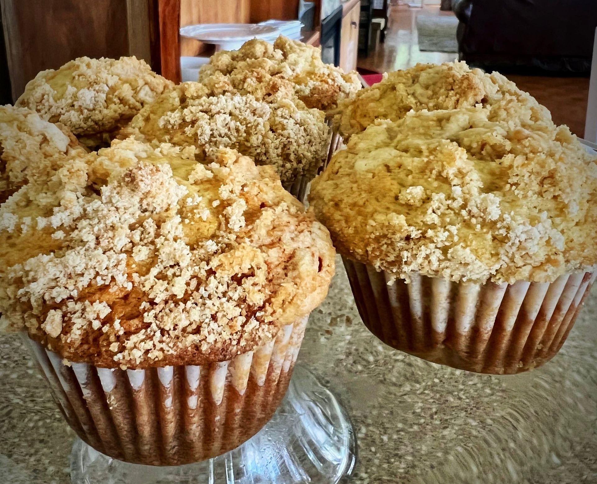 Four crumb-topped muffins on a glass stand, with a brown-and-gold color and textured tops.