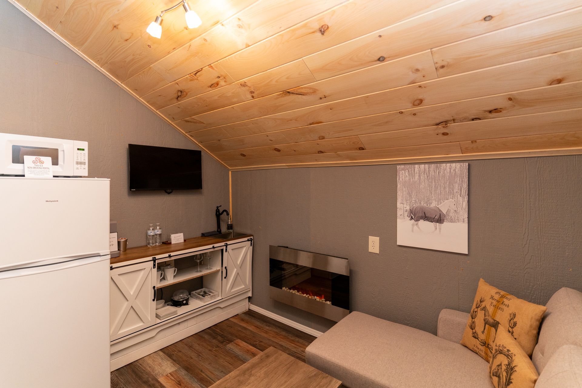 Cozy attic lounge with sloped wood ceiling, gray walls, sofa, TV, and small kitchen area