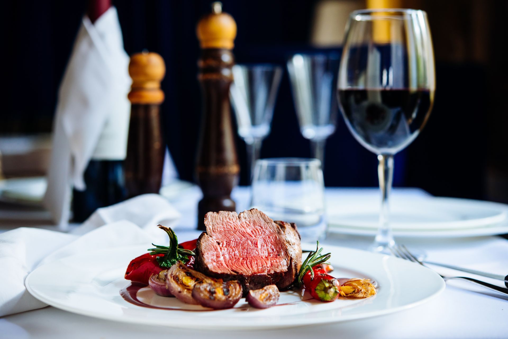 Plated steak with vegetables on a white tablecloth, with wine glass and dining setup in the background