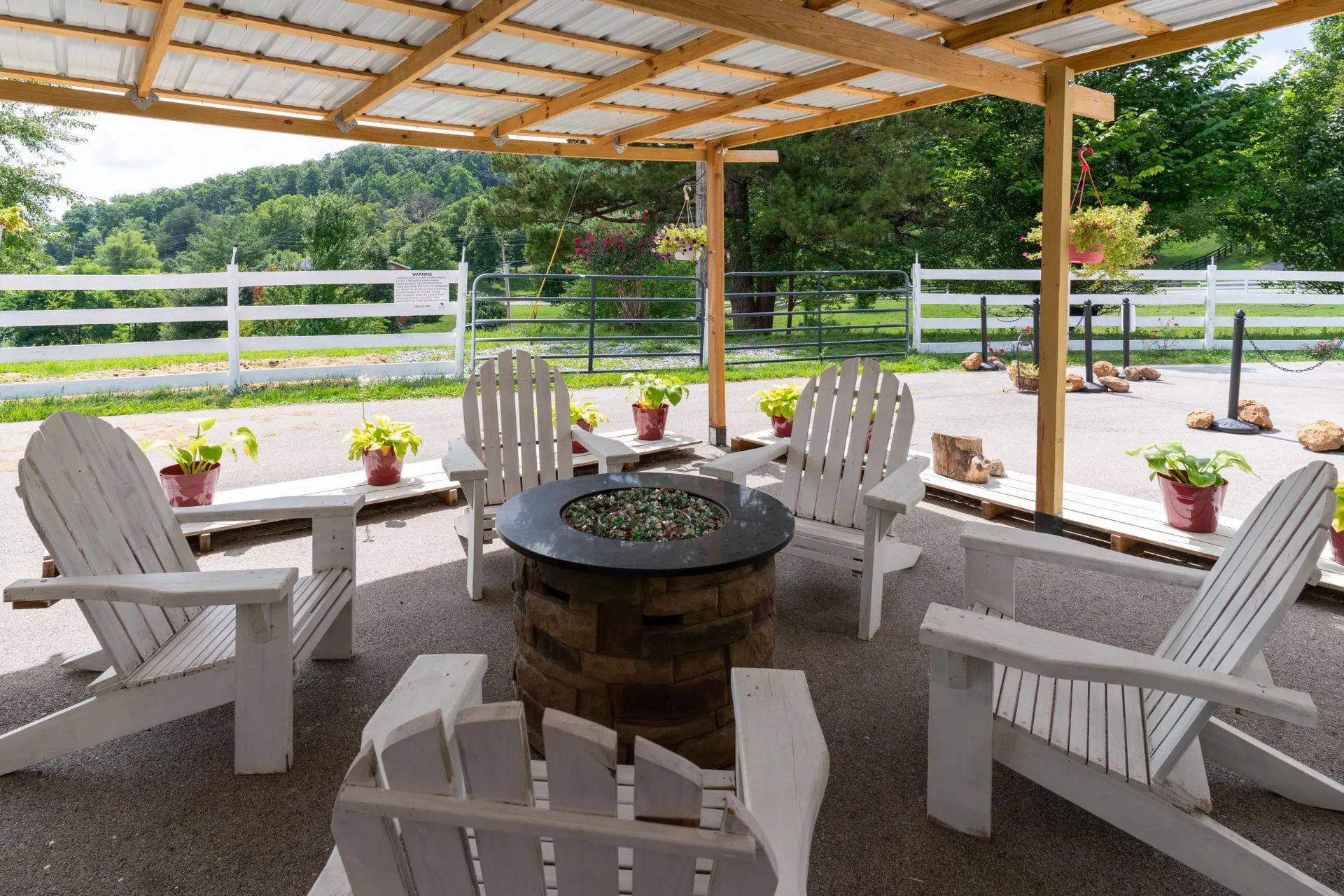 Covered patio with white Adirondack chairs around a fire pit, overlooking a grassy yard and fence.
