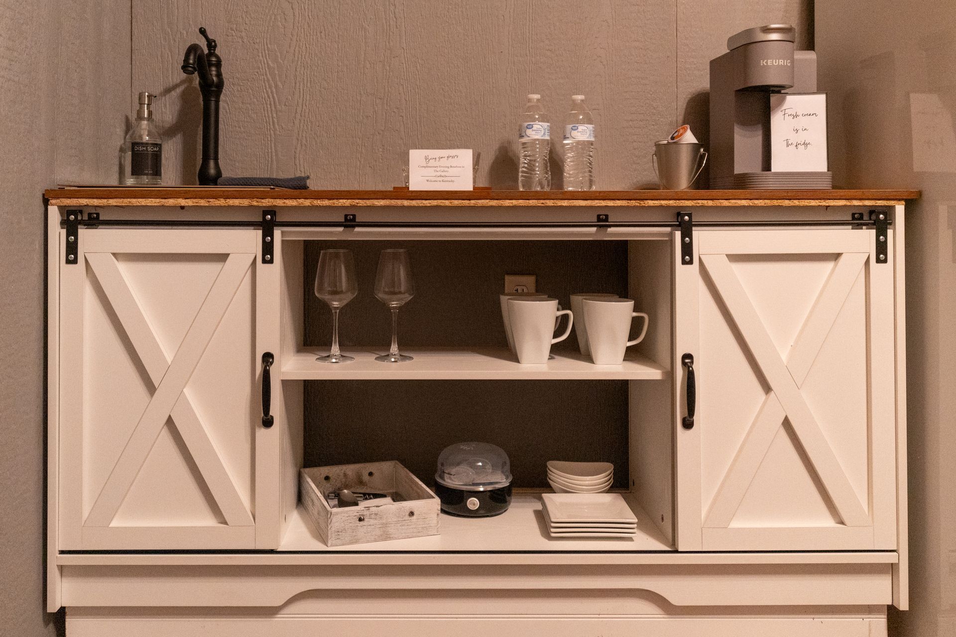White cabinet bar cart with glasses, mugs, and bottles on top against a textured wall.