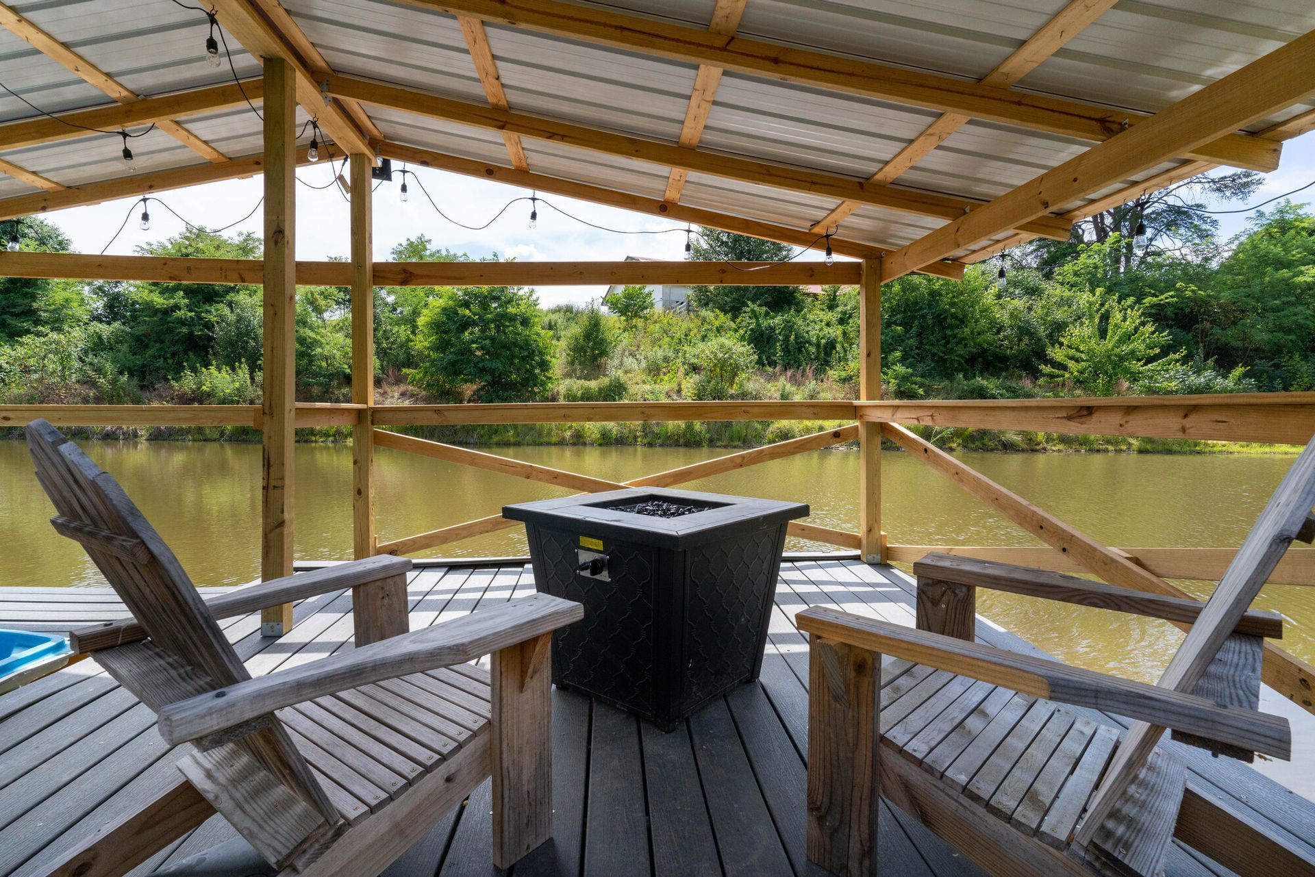 Covered wooden deck with Adirondack chairs and a black table overlooking a river and trees