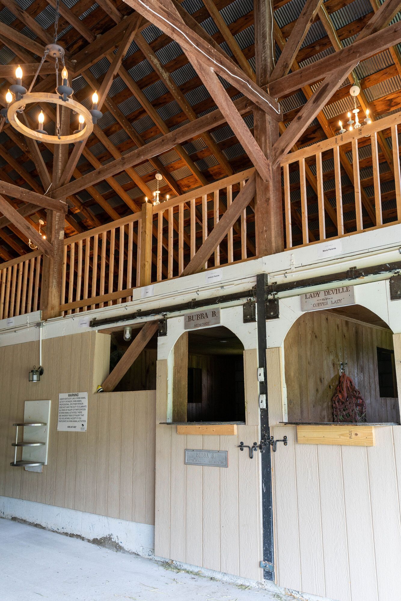 Rustic barn interior with wooden stalls, loft railings, and hanging lights under a timber roof