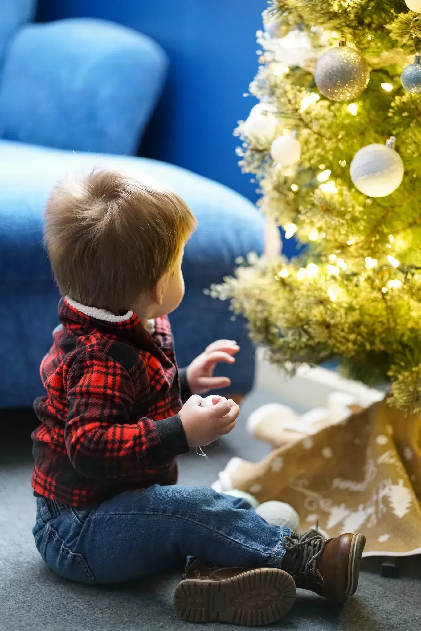 Toddler sitting by a lit Christmas tree, reaching toward ornaments on the floor