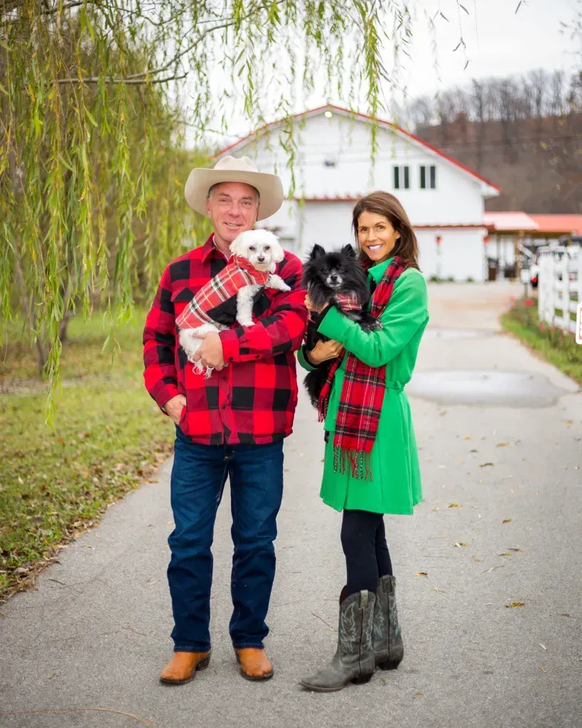 Couple holding two small dogs on a driveway near a white farmhouse, wearing red and green plaid outfits.