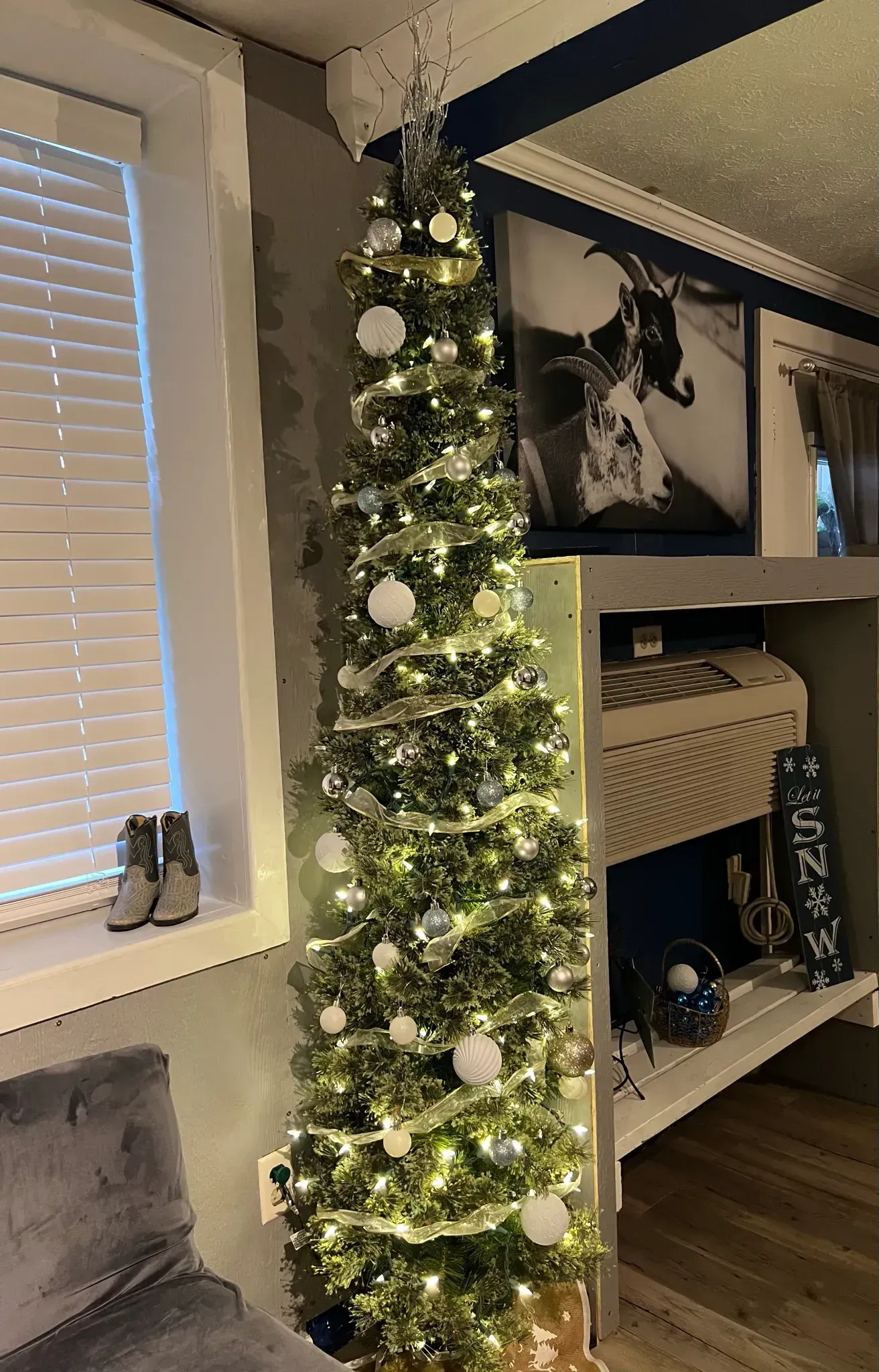 Tall decorated Christmas tree with white ornaments in a living room beside a window and fireplace bookshelf