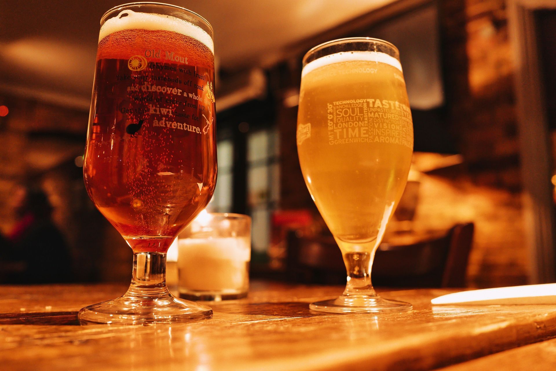 Two beer glasses on a wooden bar in warm amber light, one dark ale and one pale beer