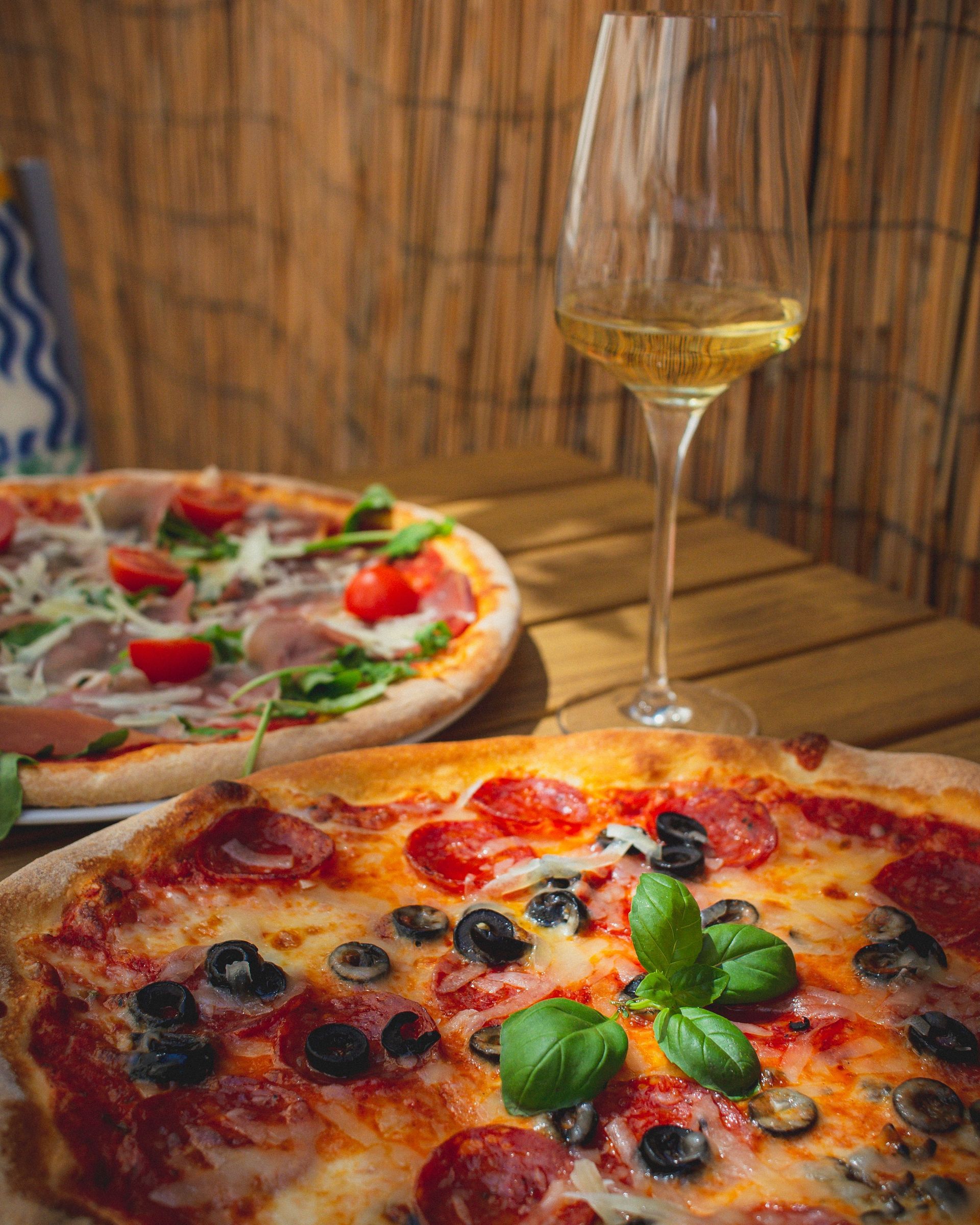 Two pizzas on a wooden table with a glass of white wine, in a rustic setting