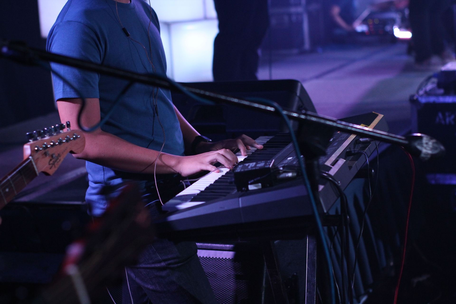 Musician playing keyboard on a dimly lit stage with purple lighting and a microphone stand nearby