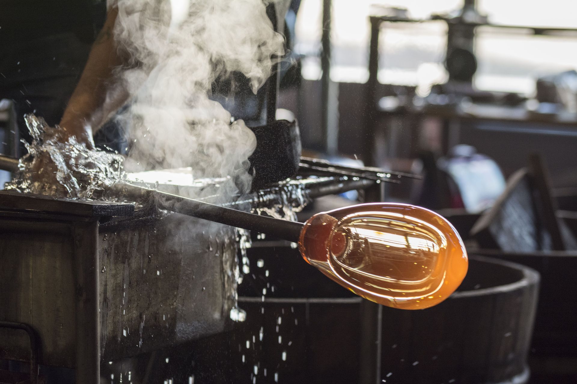Glassblower shaping a glowing orange glass piece over a steaming furnace