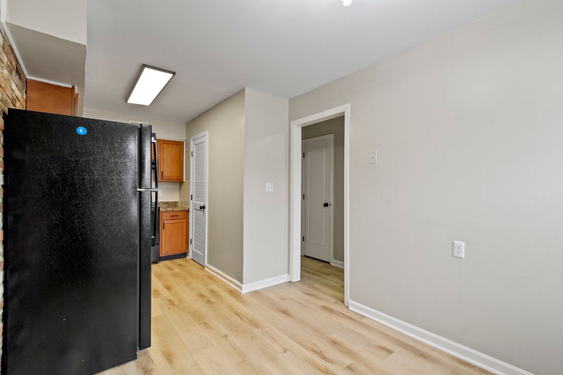 Kitchen with black fridge, wooden cabinets, and light wood flooring. A doorway and laundry-style door.