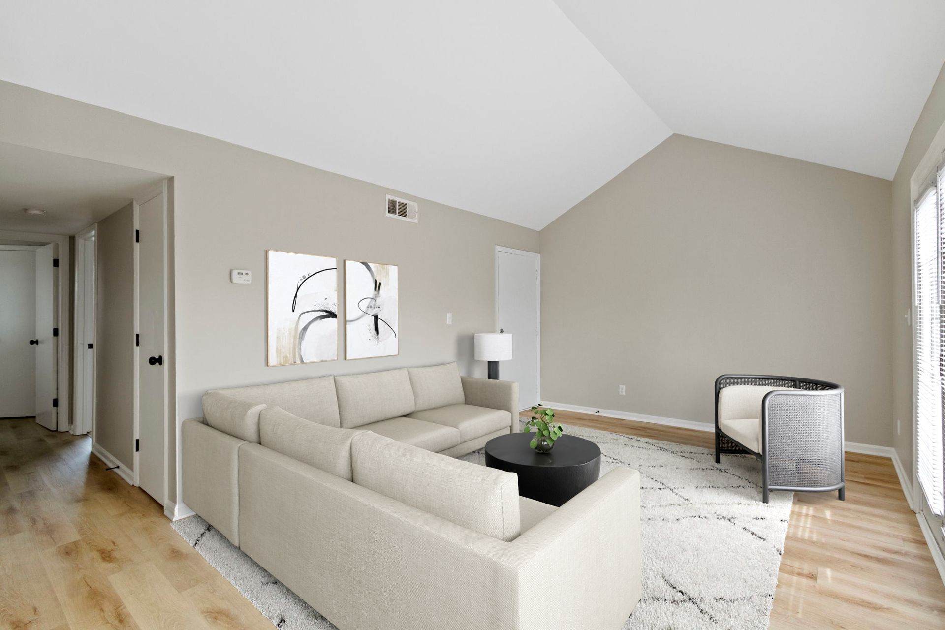 Beige living room with sectional sofa, art, round coffee table, wicker chair, and light wood floors.