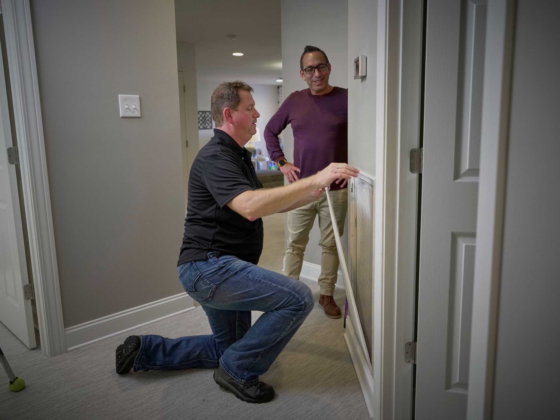 Two people installing a white safety gate in a hallway, one kneeling and one standing nearby. Two people installing a white safety gate in a hallway, one kneeling and one standing nearby.