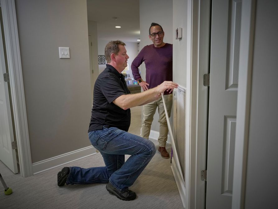Two people installing a white safety gate in a hallway, one kneeling and one standing nearby. Two people installing a white safety gate in a hallway, one kneeling and one standing nearby.