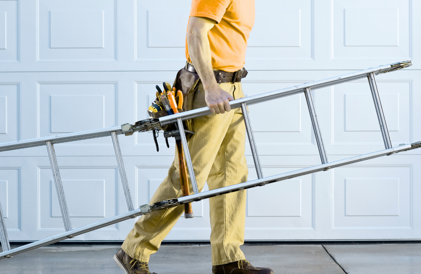 Person carrying a ladder and power tool in front of a garage door