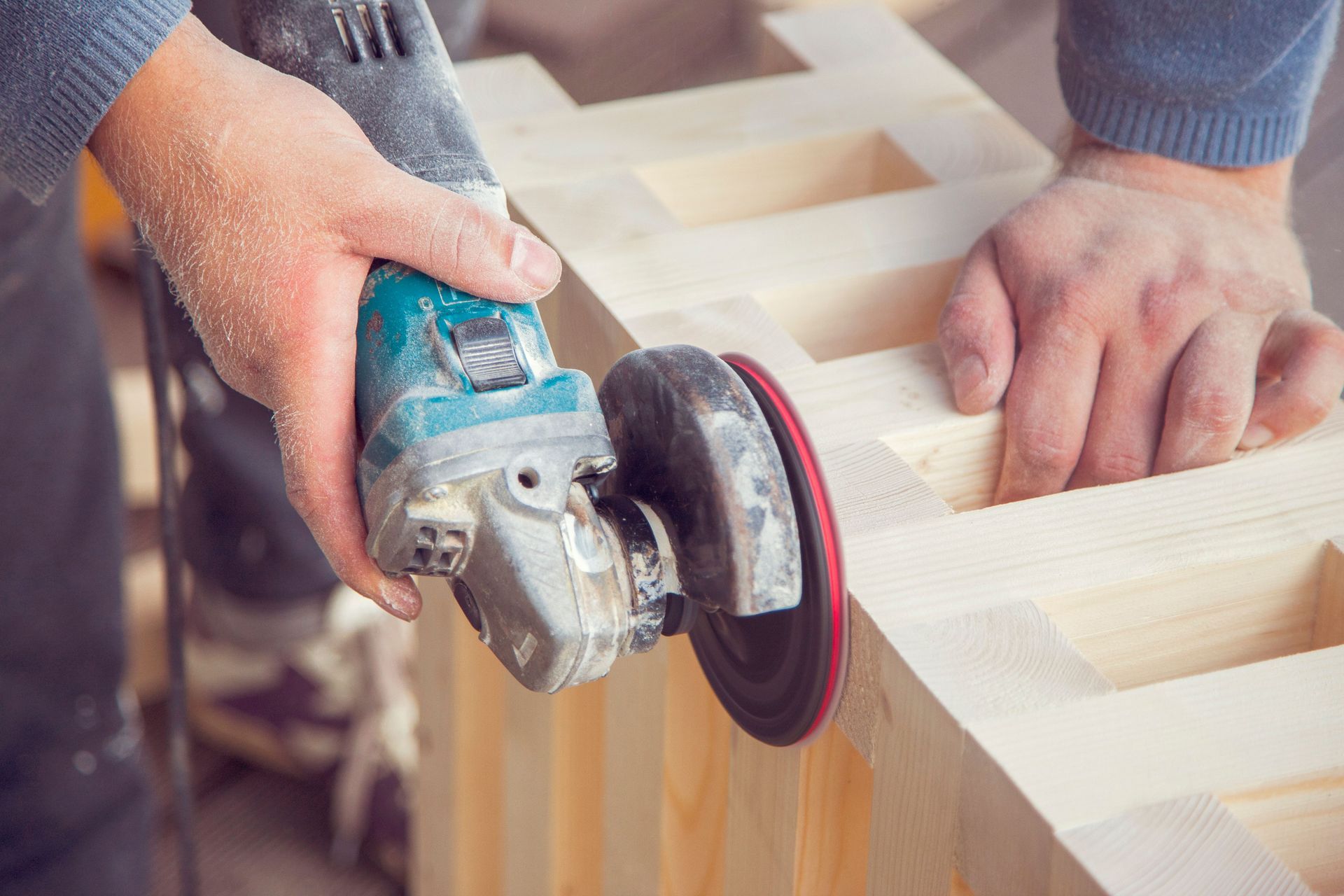Hands using an angle grinder to smooth a wooden plank in a workshop.