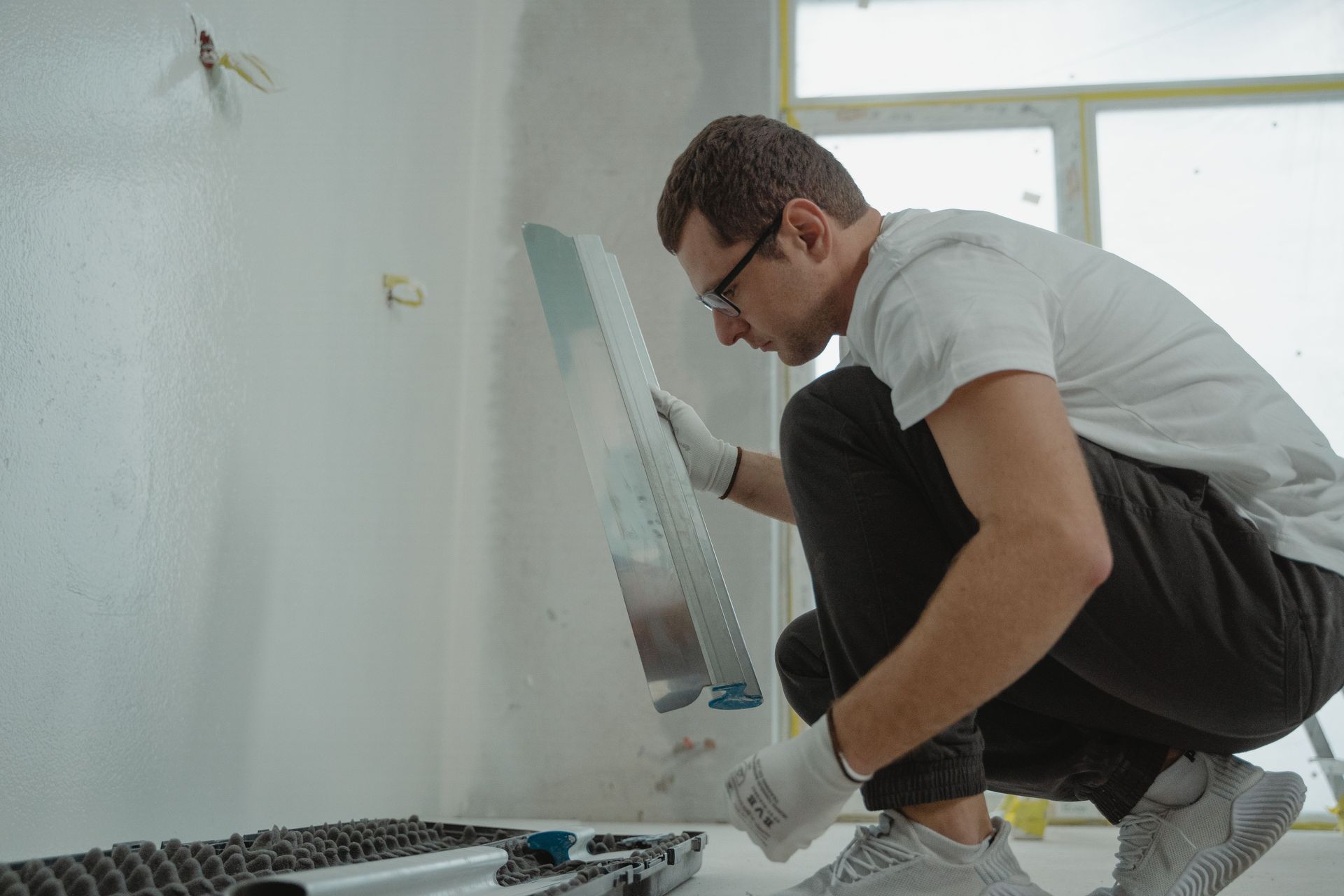 Person kneeling and painting a wall in a white room, wearing gloves and glasses