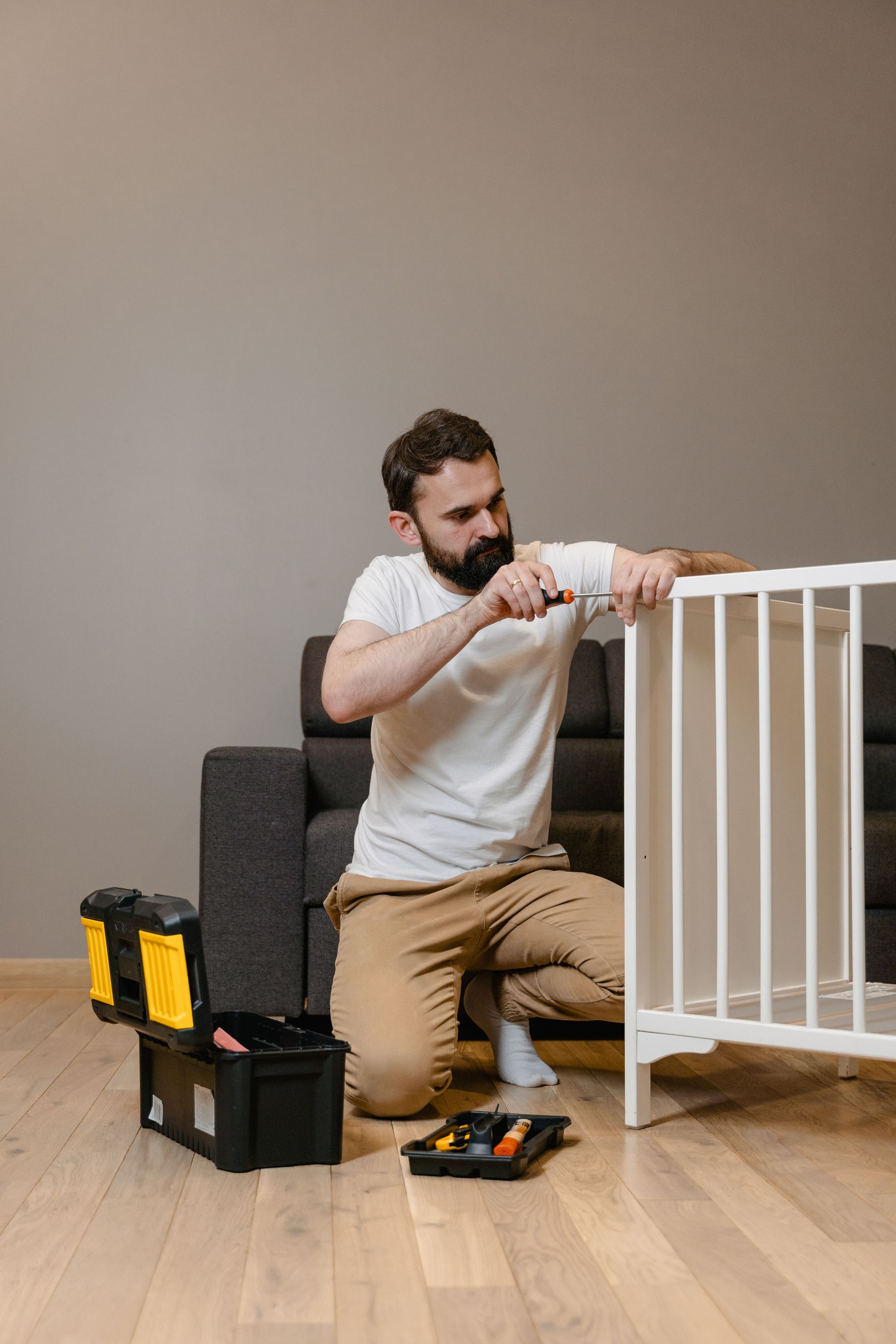 Man assembling a white baby crib with tools on a wood floor beside a gray chair.