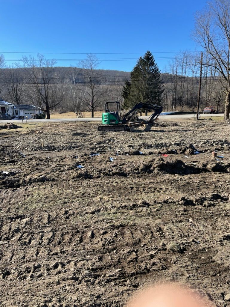 Excavator grading a muddy lot beside a road with trees and hills in the background