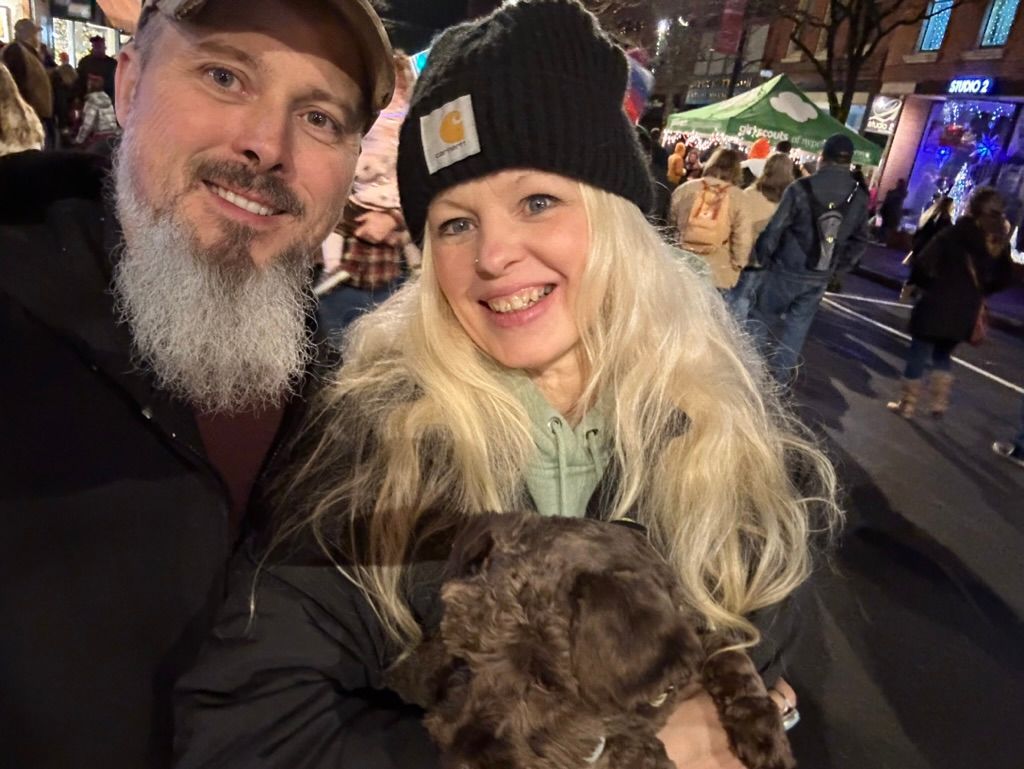 Two people smiling at a night market, holding a small dog, with colorful lights in the background