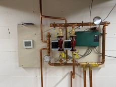 Mechanical room with copper pipes, gauges, pumps, and a wall thermostat/controller.