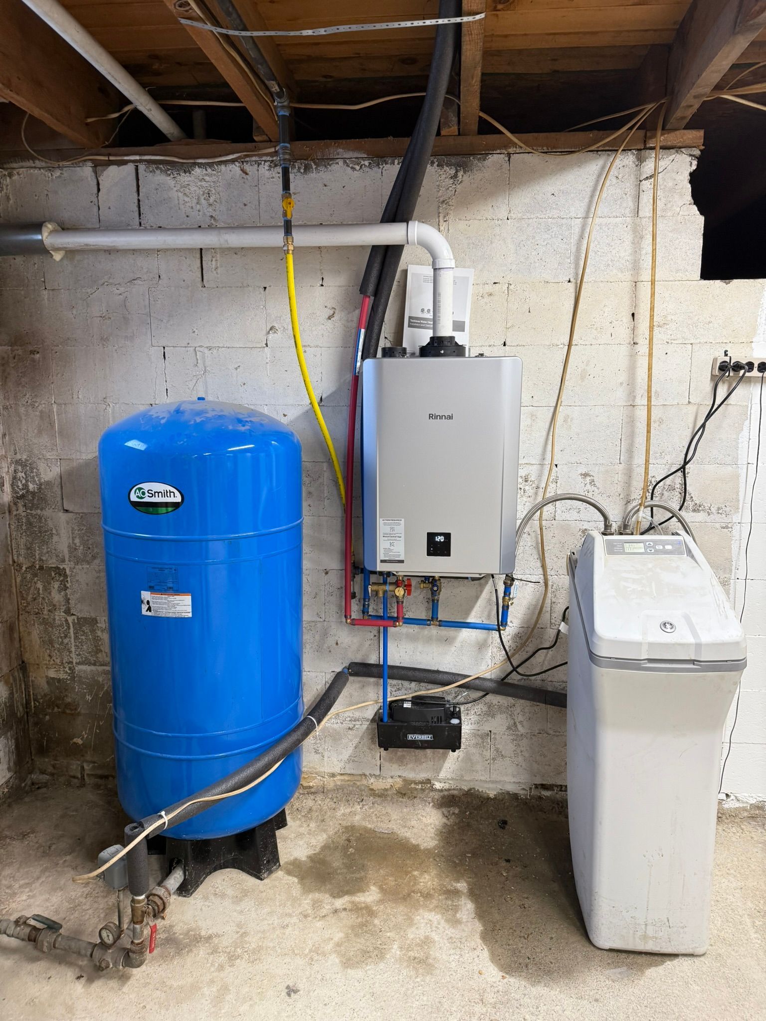 Basement utility area with blue water softener, gray heater, and white appliance beside exposed pipes and wiring