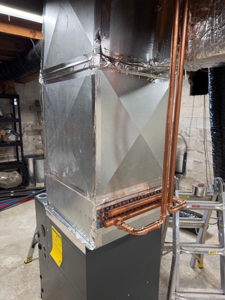 Metal HVAC ductwork in a basement utility room, with insulated silver ducts and copper pipes above a black unit.
