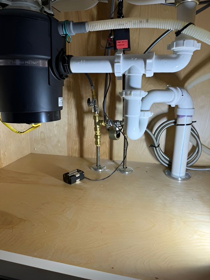Under-sink plumbing with white pipes, brass valves, and a black garbage disposal on a wooden cabinet floor
