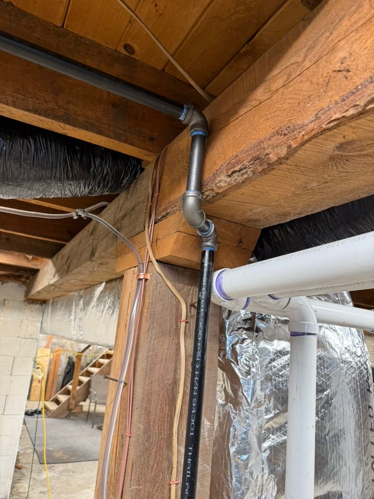 Basement ceiling with exposed wooden beams, metal ductwork, and white PVC pipes near a foil-covered wall.
