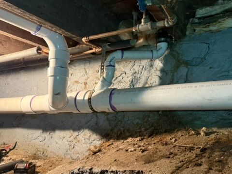 White PVC pipes and joints running along a basement wall and dirt floor.